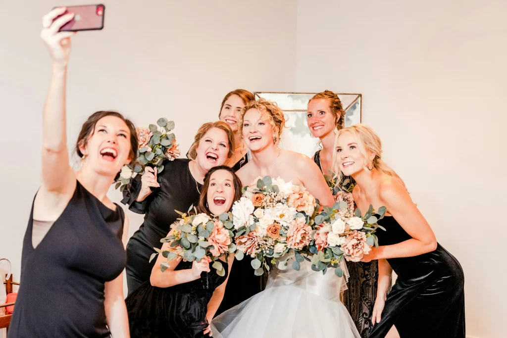 A group of bridesmaids wearing black dresses, stood with the bride taking a selfie